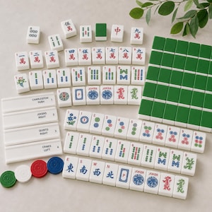 May include: A Mahjong set featuring white tiles with red, blue, green, and black characters, symbols, and dragon motifs. The set includes four white rectangular racks labelled with text, several solid green tiles, and four circular coloured tokens.
