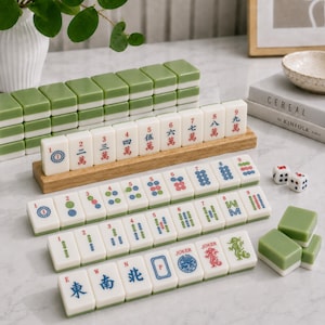 K&ouml;nnte beinhalten: Ein Mahjong-Set mit wei&szlig;en Spielsteinen mit gr&uuml;ner R&uuml;ckseite, die auf Holzst&auml;ndern und der Oberfl&auml;che angeordnet sind. Die Steine zeigen verschiedene chinesische Schriftzeichen, Zahlen und Symbole in roter, blauer und gr&uuml;ner Tinte sowie Joker-Steine und zwei W&uuml;rfel.