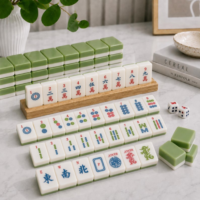 K&ouml;nnte beinhalten: Ein Mahjong-Set mit wei&szlig;en Spielsteinen mit gr&uuml;ner R&uuml;ckseite, die auf Holzst&auml;ndern und der Oberfl&auml;che angeordnet sind. Die Steine zeigen verschiedene chinesische Schriftzeichen, Zahlen und Symbole in roter, blauer und gr&uuml;ner Tinte sowie Joker-Steine und zwei W&uuml;rfel.