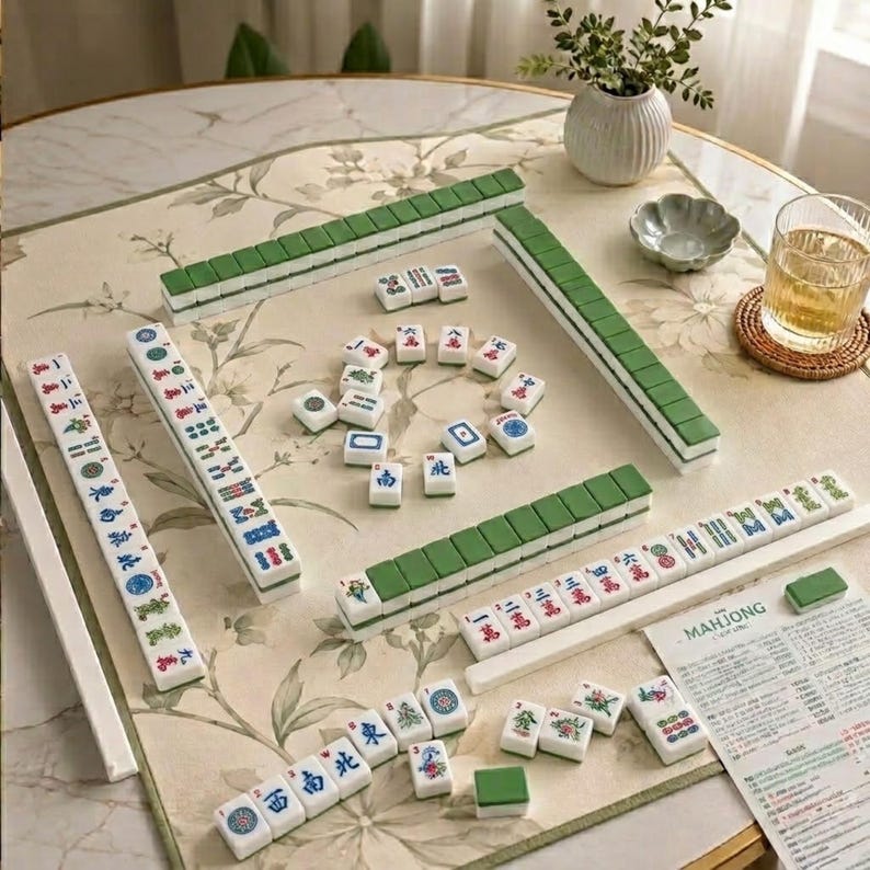 May include: A Mahjong set featuring white tiles with green backs and colourful character, bamboo, and dot markings. The set includes arranged rows, scattered tiles, and a printed instruction sheet with the text Mahjong, all displayed on a floral mat.