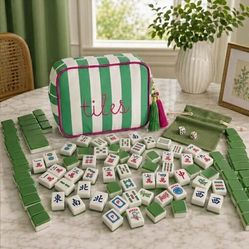 May include: A green and white striped zipped storage bag with the word tiles embroidered in pink, accompanied by a set of white mahjong tiles with various red, green, and blue characters, two dice, and a small green fabric drawstring pouch.