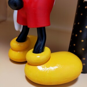 May include: Close-up of a wooden Mickey Mouse figurine. The figure features bright yellow shoes, red shorts, black legs, and a black tail. The background is a neutral beige.