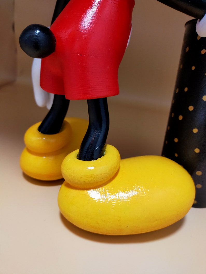 May include: Close-up of a wooden Mickey Mouse figurine. The figure features bright yellow shoes, red shorts, black legs, and a black tail. The background is a neutral beige.