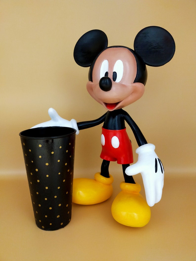 May include: A Mickey Mouse figurine with a black body, red shorts, and yellow shoes stands next to a black cylindrical container with gold polka dots. The figurine has white gloves and a tan face.