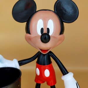 May include: A Mickey Mouse figurine with black ears, a black body, red shorts with white buttons, and yellow shoes. The figure has white gloves and a smiling expression. It is holding a black container with gold dots.