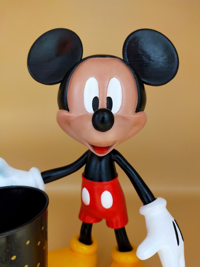 May include: A Mickey Mouse figurine with black ears, a black body, red shorts with white buttons, and yellow shoes. The figure has white gloves and a smiling expression. It is holding a black container with gold dots.