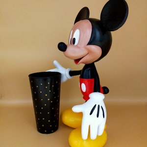 May include: A Mickey Mouse figurine with a black body, red shorts, and yellow feet. The figure is holding a black cup with gold polka dots. The background is a solid tan color.