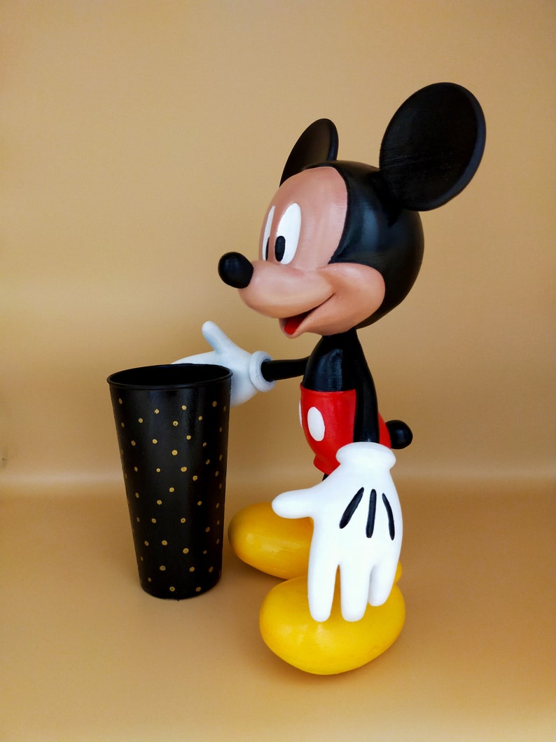 May include: A Mickey Mouse figurine with a black body, red shorts, and yellow feet. The figure is holding a black cup with gold polka dots. The background is a solid tan color.