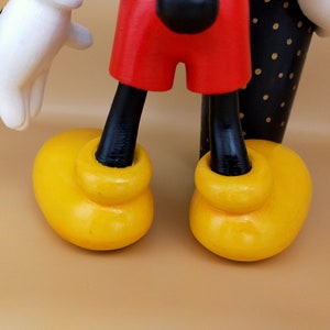 May include: Close-up of a wooden Mickey Mouse figure. The figure has yellow shoes, red shorts, black legs, a white glove, and a black and white polka-dotted arm. The background is a solid tan color.