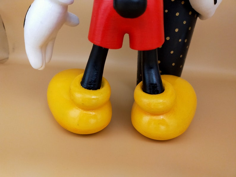 May include: Close-up of a wooden Mickey Mouse figure. The figure has yellow shoes, red shorts, black legs, a white glove, and a black and white polka-dotted arm. The background is a solid tan color.
