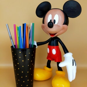 May include: A Mickey Mouse figurine stands next to a black pen holder with gold polka dots. The pen holder contains colorful pens and straws. Mickey Mouse is wearing red shorts, yellow shoes, and white gloves.