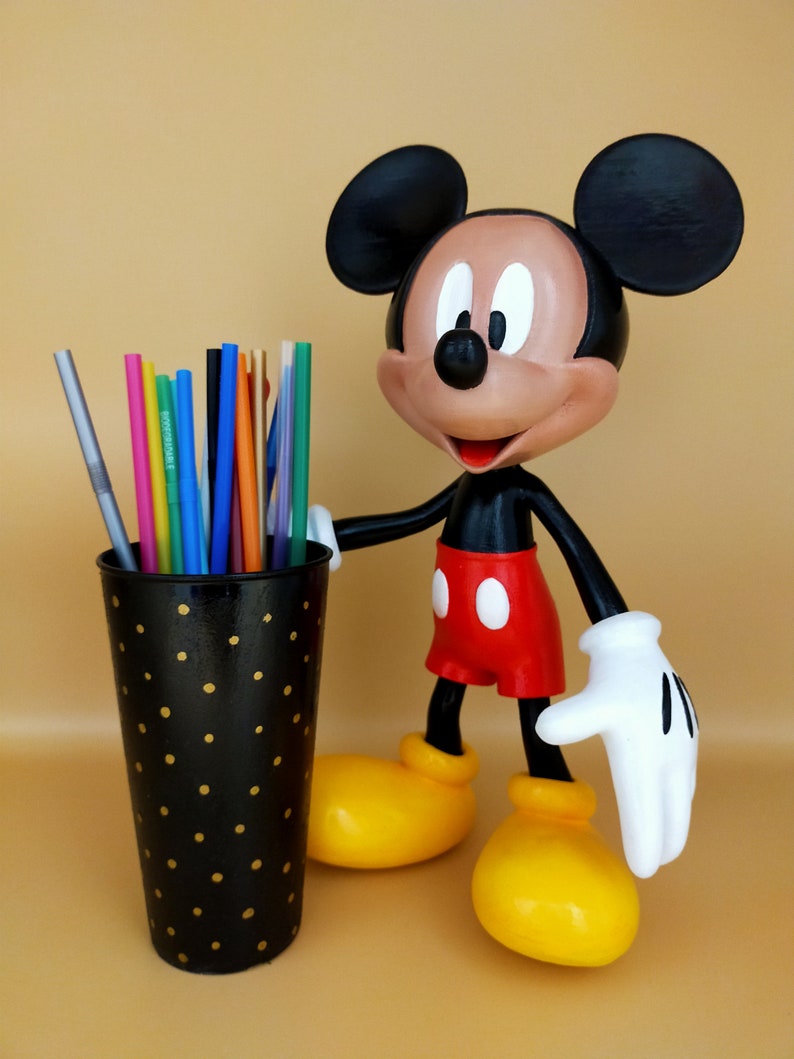 May include: A Mickey Mouse figurine stands next to a black pen holder with gold polka dots. The pen holder contains colorful pens and straws. Mickey Mouse is wearing red shorts, yellow shoes, and white gloves.