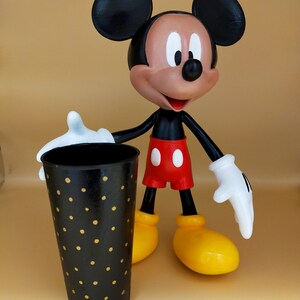 May include: A Mickey Mouse figurine stands next to a black tumbler with gold polka dots. The figurine has black ears, a brown face, red shorts, yellow shoes, and white gloves. The tumbler is cylindrical and appears to be made of metal.