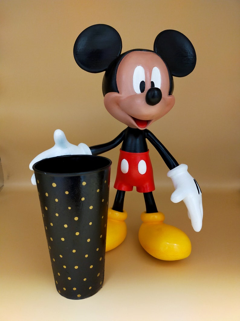 May include: A Mickey Mouse figurine stands next to a black tumbler with gold polka dots. The figurine has black ears, a brown face, red shorts, yellow shoes, and white gloves. The tumbler is cylindrical and appears to be made of metal.
