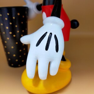 May include: A figurine of a cartoon character with white gloves, a red shirt, and yellow shoes. The black and white polka-dotted cup is in the background. The character's white glove has black lines on the fingers.