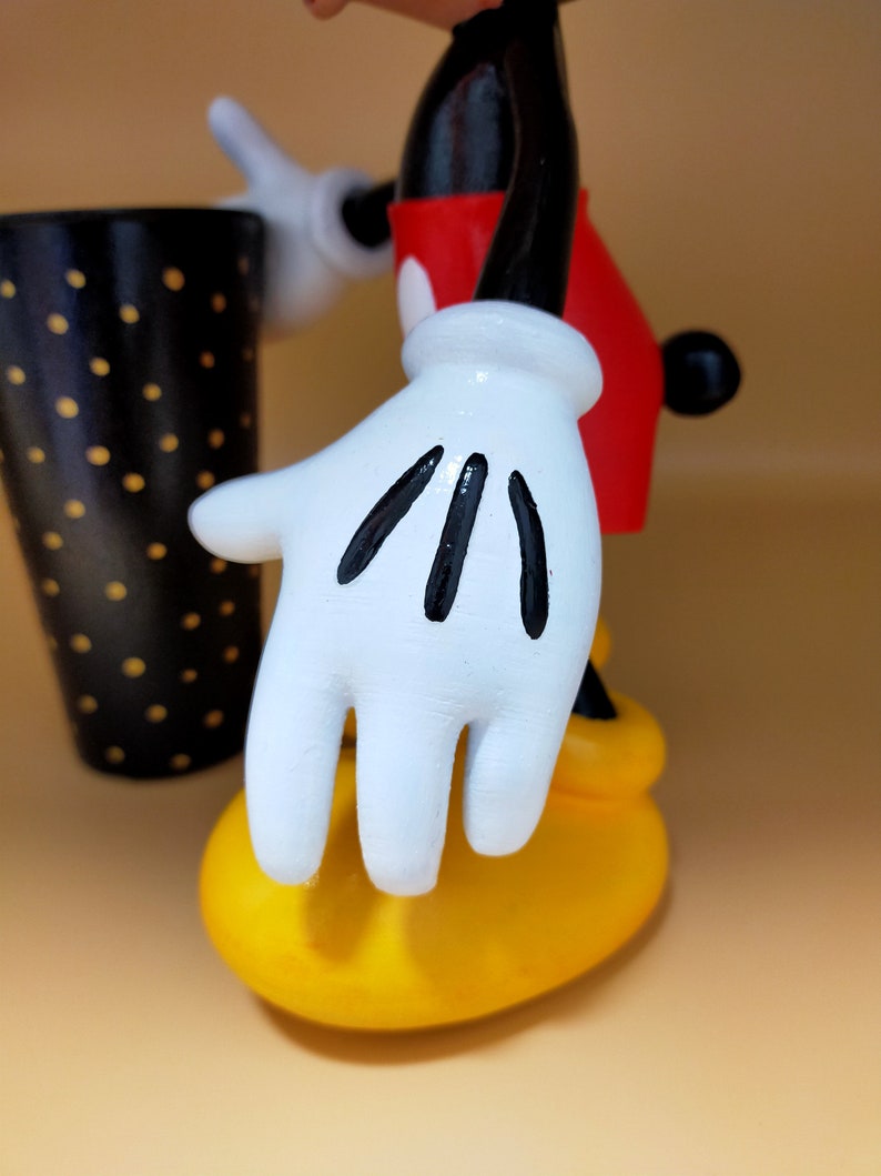 May include: A figurine of a cartoon character with white gloves, a red shirt, and yellow shoes. The black and white polka-dotted cup is in the background. The character's white glove has black lines on the fingers.