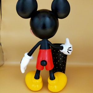 May include: A Mickey Mouse figurine, viewed from the back, with black ears, head, and arms. The figure wears red shorts, yellow shoes, and white gloves. A black and gold polka-dot cup is next to the figure.