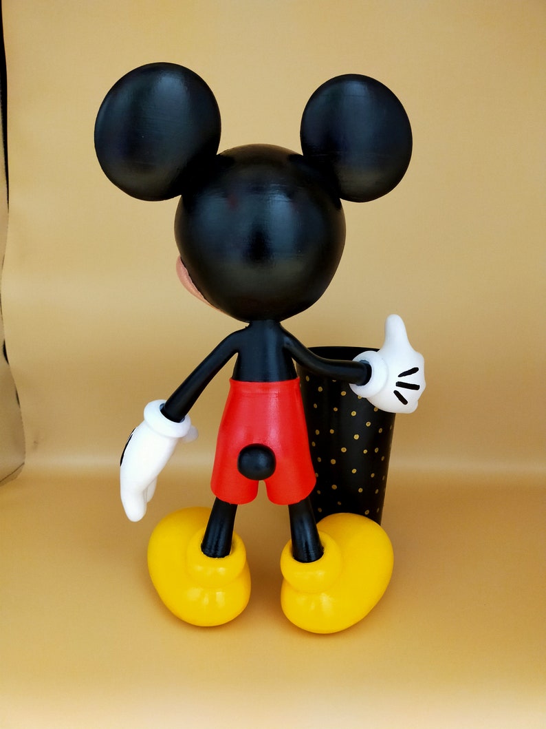 May include: A Mickey Mouse figurine, viewed from the back, with black ears, head, and arms. The figure wears red shorts, yellow shoes, and white gloves. A black and gold polka-dot cup is next to the figure.