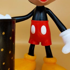 May include: A close-up of a Mickey Mouse figurine. The figure has a black torso and arms, red shorts with white buttons, yellow shoes, and white gloves. The figurine is holding a black mug with yellow polka dots.