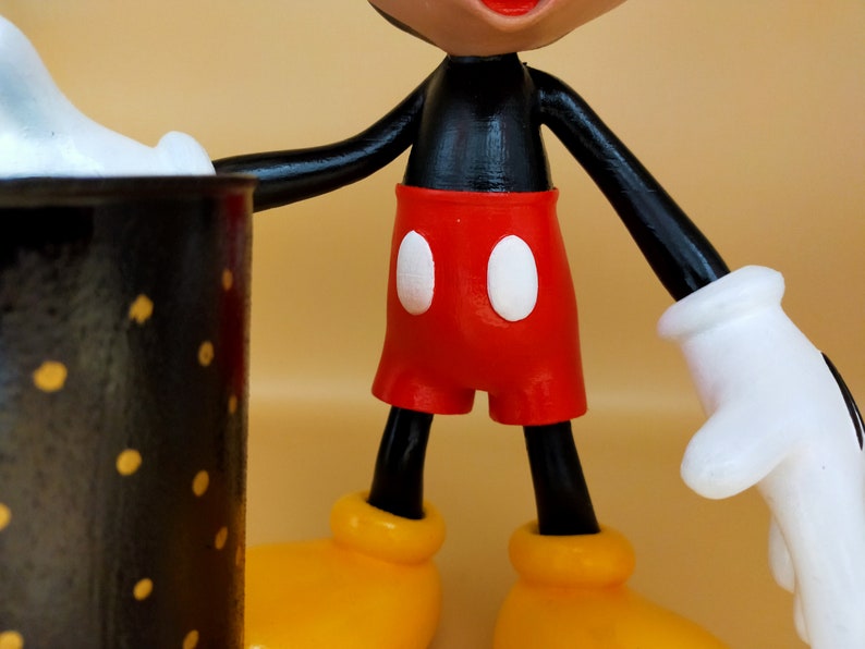 May include: A close-up of a Mickey Mouse figurine. The figure has a black torso and arms, red shorts with white buttons, yellow shoes, and white gloves. The figurine is holding a black mug with yellow polka dots.