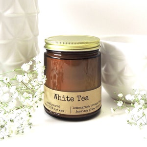 May include: A brown glass candle jar with a gold lid. The label reads "White Tea" and lists the scent notes: lemongrass, orange, jasmine, plum, cedar. The candle is hand-poured with 100% soy wax.
