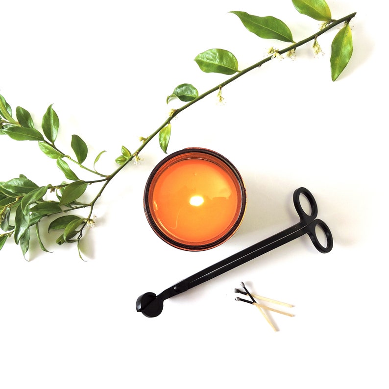 May include: A lit amber glass candle with a wick burning in the center. A black candle snuffer with a rounded handle and a pair of scissor-like prongs is lying on a white surface. Three used matches are also on the surface.