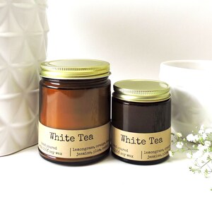 May include: Two amber glass candle jars with gold lids. The labels are brown and say "White Tea" with the scent notes listed below. The larger jar is in the foreground and the smaller jar is behind it.
