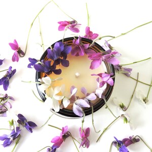 May include: A glass candle jar with a light brown candle surrounded by a ring of purple, pink, and white flowers. The flowers are arranged in a circle around the candle, and the jar is sitting on a white surface.