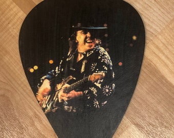 May include: A black and white guitar pick with a photo of a musician playing a guitar on stage. The musician is wearing a black hat and a patterned shirt.