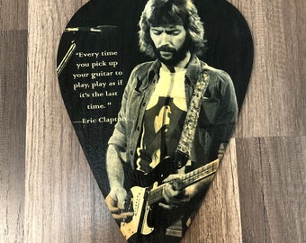 May include: A black guitar pick with a photo of Eric Clapton playing a guitar. The pick has a quote from Eric Clapton that reads "Every time you pick up your guitar to play, play as if it's the last time."
