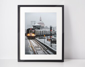 May include: A black framed photograph of a train traveling on a snowy track in front of a building with a dome. The train is facing the camera and the building is in the background.
