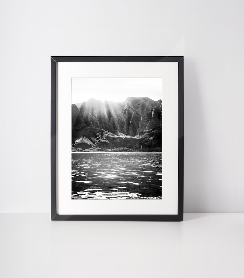 May include: Black and white photograph of a mountain range with sunbeams shining through the peaks, reflecting on the calm water below.