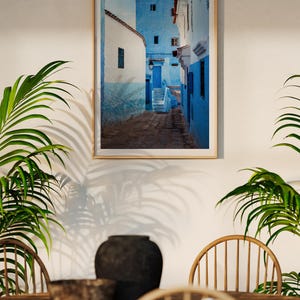 May include: A framed photograph of a narrow, blue-painted alleyway in a Moroccan city. The alleyway is lined with white-washed buildings and has a stone floor. The photograph is in a wooden frame.
