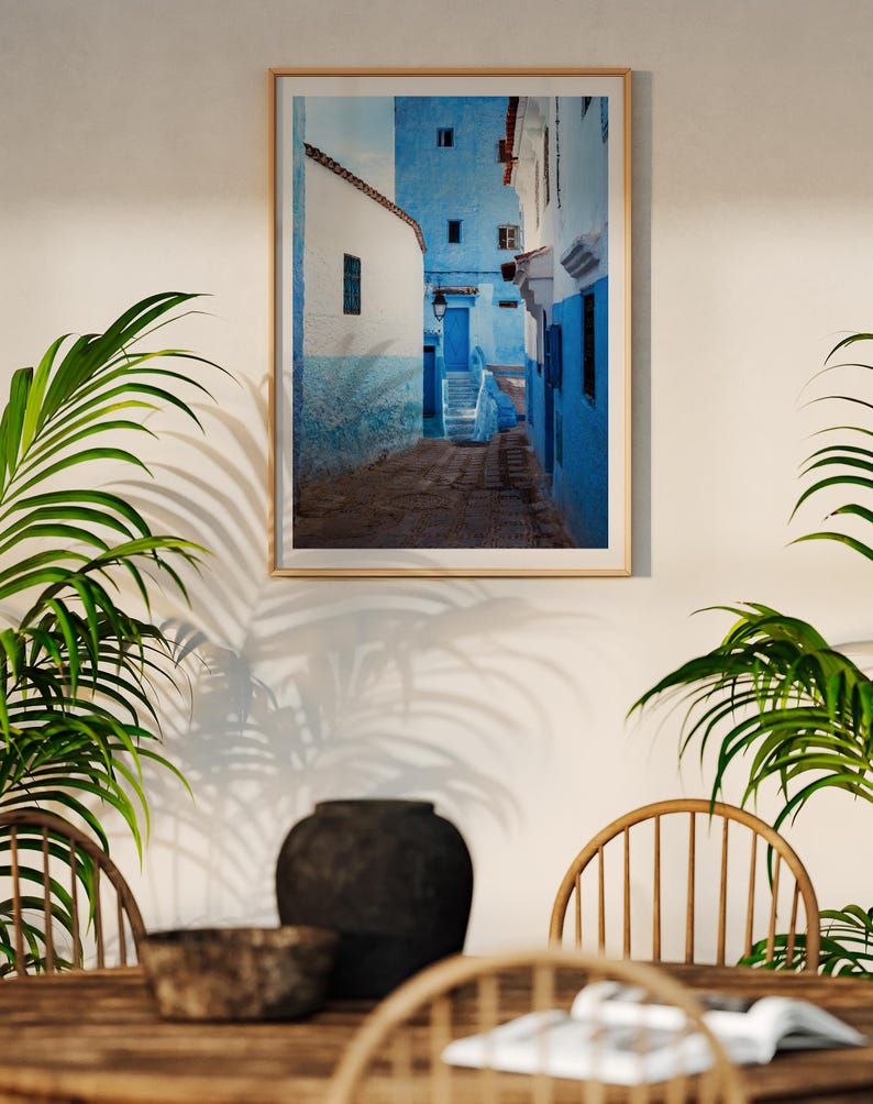 May include: A framed photograph of a narrow, blue-painted alleyway in a Moroccan city. The alleyway is lined with white-washed buildings and has a stone floor. The photograph is in a wooden frame.