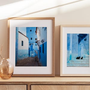 May include: Two framed photographs of a blue and white Moroccan street scene. The first photo shows a narrow street with blue buildings and a staircase. The second photo shows a close-up of a blue wall with a staircase and two black cats sitting on the steps.