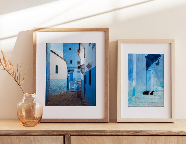 May include: Two framed photographs of a blue and white Moroccan street scene. The first photo shows a narrow street with blue buildings and a staircase. The second photo shows a close-up of a blue wall with a staircase and two black cats sitting on the steps.