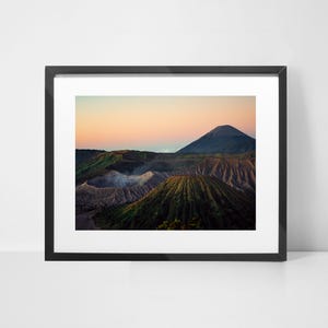 Op de afbeelding: Een panoramisch uitzicht op een vulkanisch landschap in Indonesië. De afbeelding toont een reeks vulkanische toppen, waaronder een grote, slapende vulkaan op de achtergrond. De lucht is zacht roze en oranje, wat duidt op een zonsopgang of zonsondergang. De voorgrond is bedekt met vulkanische as en vegetatie.