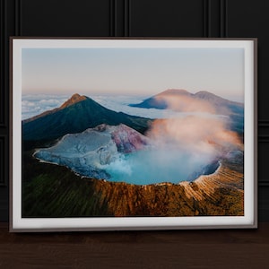 Könnte beinhalten: Gerahmter Landschaftsdruck mit einem Vulkankrater mit einem türkisfarbenen See, umgeben von Bergen und Wolken. Das Bild zeigt eine lebendige Farbpalette aus Grün-, Braun-, Blau- und Rosatönen und erzeugt eine ruhige und natürliche Szene.