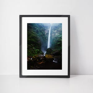 May include: A framed photograph of a waterfall cascading down a cliff face. Lush green foliage surrounds the falls, with rocks and a small stream at the base. The image is framed in a black border.
