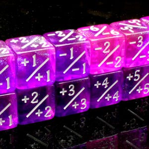 May include: A set of twelve translucent purple six-sided dice arranged in two rows. Each die face features white engraved numbers separated by a diagonal slash, displaying various positive and negative integer combinations ranging from -6 to +6.