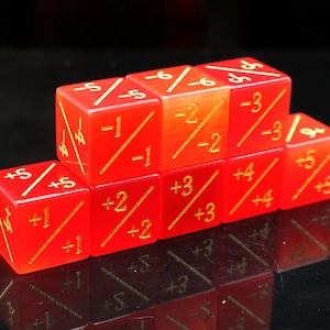 May include: A collection of seven red dice with gold markings. Each die features numbers and symbols, including plus and minus signs, and diagonal lines. The dice are arranged in a stepped formation, with a reflective surface below.
