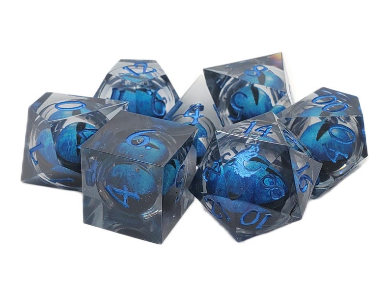 May include: Set of seven clear resin dice with blue and silver accents. Each die features blue internal designs and blue numbering. The dice are in various polyhedral shapes, including a cube, a d4, a d6, a d8, a d10, a d12, and a d20.