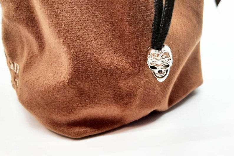 May include: A brown velvet dice bag with a silver skull charm. The bag has a drawstring closure and is perfect for storing dice.