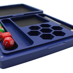 May include: A blue dice tray with a black interior, holding several dice. The tray has compartments for dice and a pen. Red and green dice are visible, along with a pen. The tray is designed for tabletop gaming.