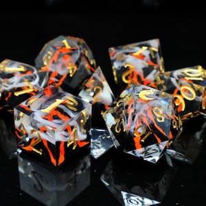 May include: A set of seven polyhedral dice with a unique design. The dice are clear with black, orange, and white inclusions, and gold-colored numbers. The dice are arranged on a reflective black surface.