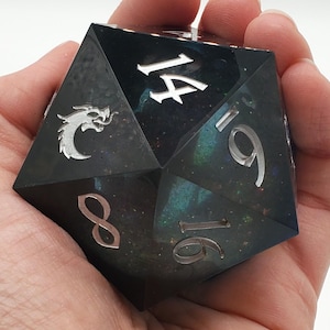 May include: A large, multi-sided die with a dark, iridescent finish, held in a hand. The die features silver numerals and a dragon emblem. The numbers 8, 9, 14, and 16 are visible. This item is likely used for tabletop gaming.