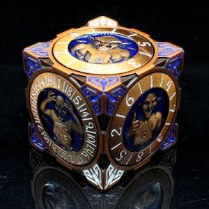 May include: A metallic bronze and blue cubic dice box featuring intricate engravings and circular insets on each face. Each side displays a sculpted goblin figure holding a die, surrounded by a numbered ring with digits ranging from 1 to 20.