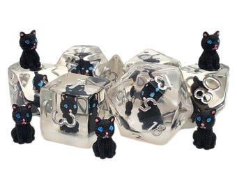 Könnte beinhalten: Satz aus transparenten, vielseitigen Würfeln mit silbernen Zahlen und schwarzen Katzenfiguren im Inneren. Kleine schwarze Katzenfiguren mit blauen Augen sind um die Würfel herum platziert. Für Tischspiele.