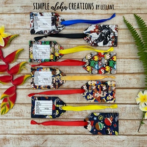 May include: Seven colorful luggage tags with superhero characters printed on them. Each tag has a white label with lines for writing personal information. The tags are attached to colorful ribbons.