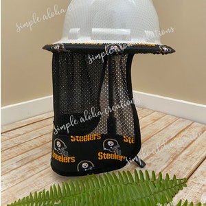 Pu&ograve; includere: Un casco bianco con un parasole in rete nera. Il parasole ha un motivo con il logo degli Steelers. Il casco &egrave; appoggiato su una superficie di legno con una pianta di felce verde in primo piano. Il testo "Neck Shade Style # 1 Side View" &egrave; visibile nell'immagine.